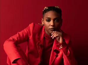 Studio portrait of Bia Ferreira dressed in shades of red, seated against a matching red background with one hand resting beneath the chin.