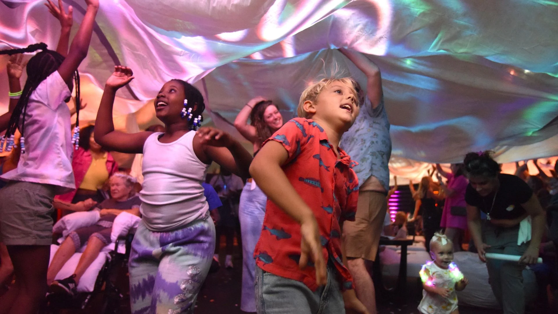 Children and families dance beneath a billowing fabric canopy illuminated with colorful lights at a lively block party.