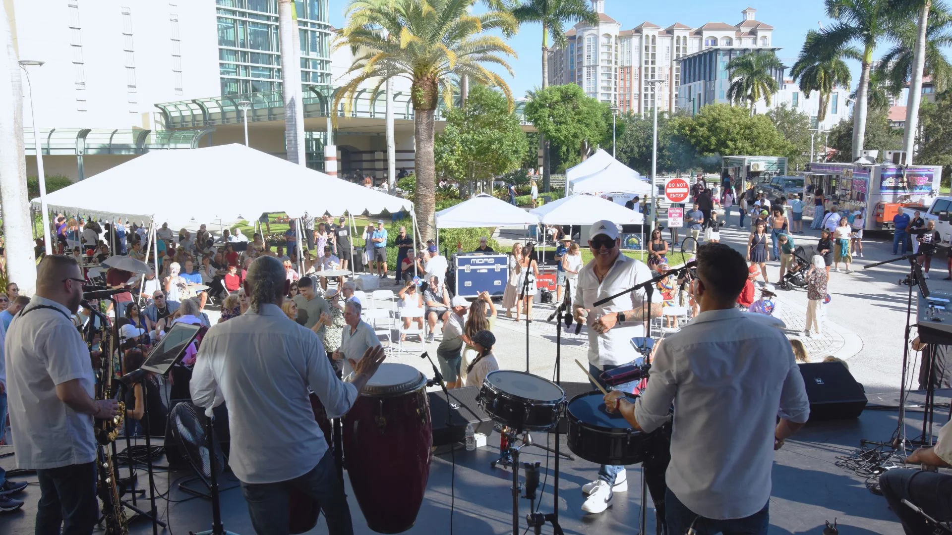 Musicians perform onstage at a sunny outdoor block party as guests gather under tents, visit food trucks, and enjoy live entertainment in a palm-lined plaza.