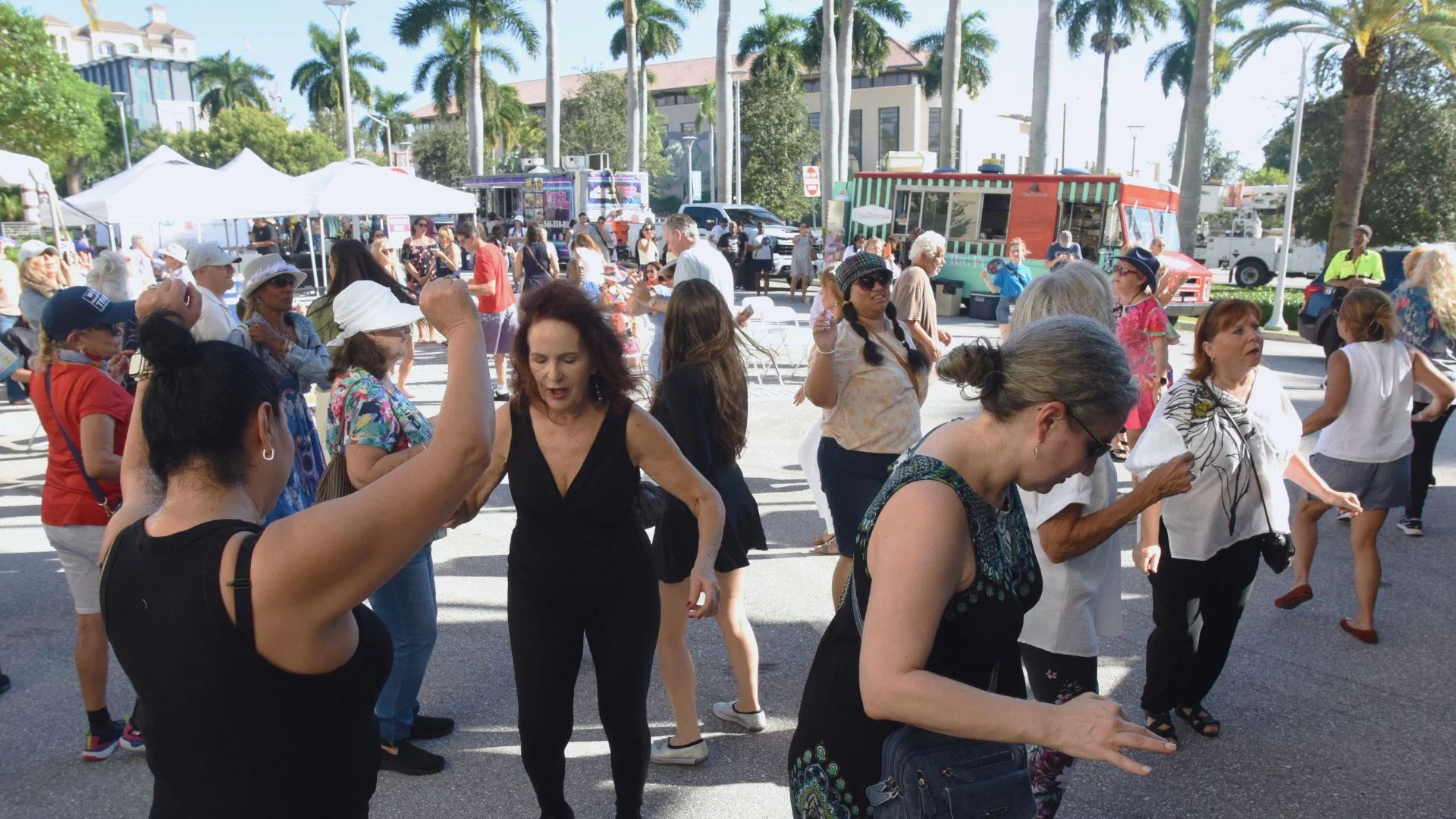 Guests dance and socialize at a sunny outdoor block party surrounded by food trucks, vendor tents, and palm trees.
