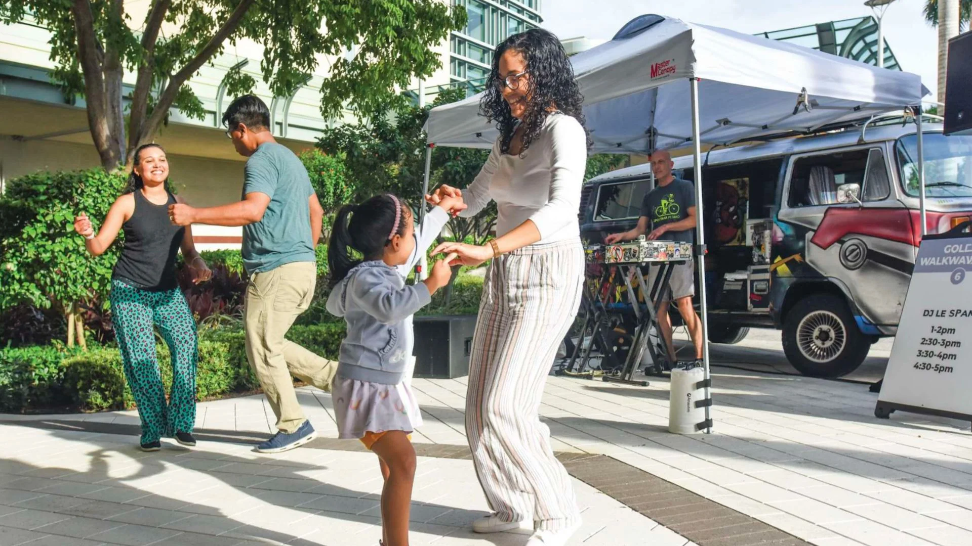Guests of all ages dance together at a sunny outdoor block party while a DJ performs under a tent in a plaza surrounded by trees and buildings.