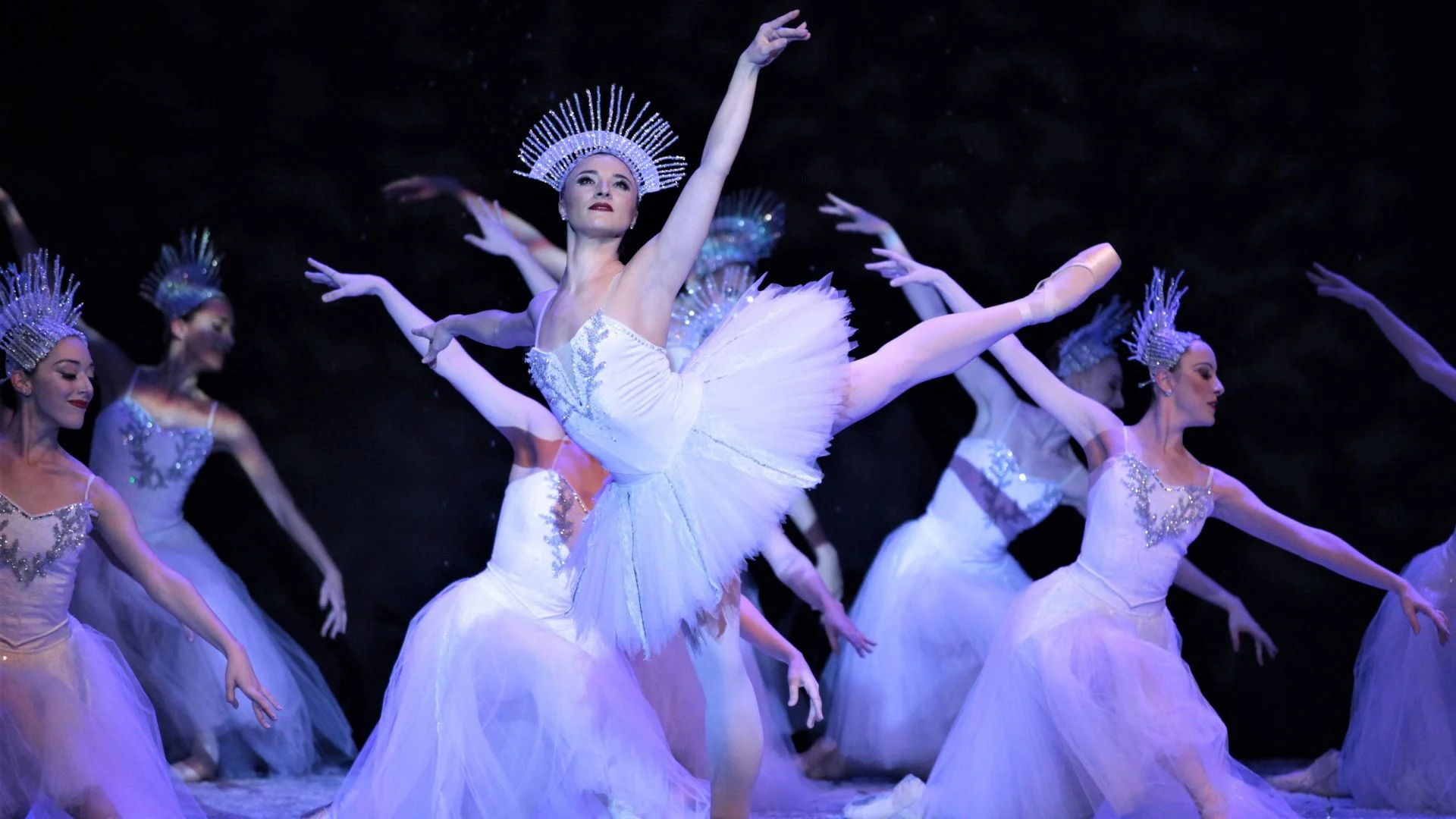 Ballet dancers in shimmering white costumes and jeweled headdresses perform a graceful ensemble piece under blue stage lighting.