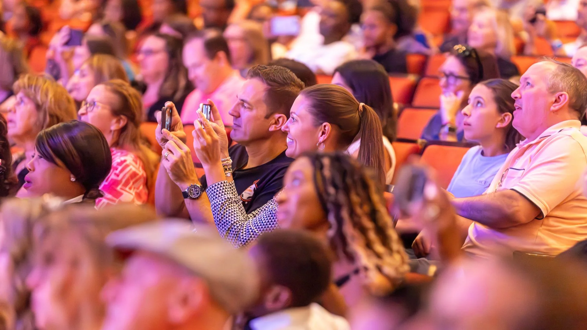 Theatergoers watch a live performance, with several audience members taking photos from their seats in a packed auditorium.