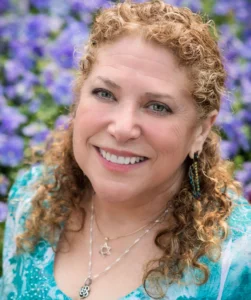 Portrait of Carrie Ayvar smiling outdoors, framed by a background of purple flowers and greenery.