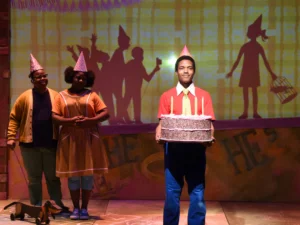 A performer wearing a party hat carries a large birthday cake with lit candles while two other performers stand nearby. Colorful silhouettes and illustrations are projected onto the backdrop of the stage.