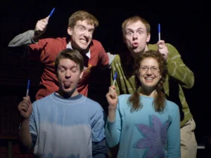 Four performers pose onstage holding pens, with playful expressions and dramatic lighting that highlights the comedic scene.