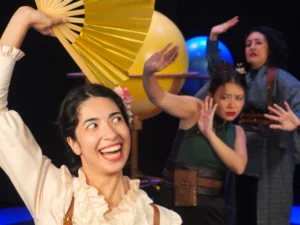 Three performers appear onstage in front of large planet props. One performer in the foreground holds an open gold fan while two others pose behind them, creating a scene inspired by storytelling and movement.