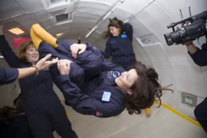 Participants wearing blue flight suits float inside a microgravity aircraft cabin while a camera operator records the activity. 