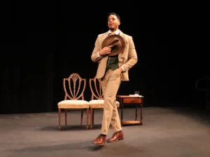 A performer in a tan suit walks across a minimally furnished stage holding a hat. Several vintage-style chairs and tables are arranged around the set for a theatrical production.