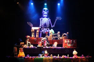 A theatrical set inspired by Día de los Muertos features a large skeleton figure towering above colorful altars, puppets, flowers, candles and decorative props on a darkened stage.