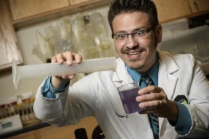 A science presenter in a laboratory coat pours liquid from a large syringe into a clear container filled with purple liquid during a hands-on science demonstration.