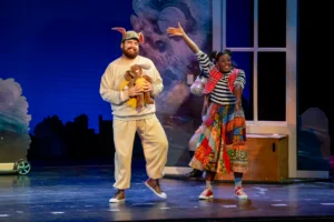 Two performers stand onstage in front of a house-like set piece. One performer wears rabbit ears and holds a stuffed rabbit while the other gestures toward the audience during a theatrical scene.