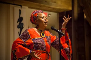 A performer wearing a brightly patterned red outfit and matching headwrap stands at a microphone, gesturing while speaking in an indoor performance space.
