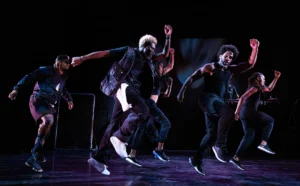 A group of dancers dressed in black leap across a stage in a synchronized performance under dramatic theatrical lighting.