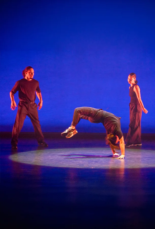3 dancers performing on stage wearing black, two standing and one doing a backbend.