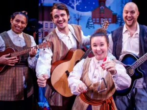Four performers in folk-style costumes play string instruments and percussion on a colorful winter-themed stage decorated with illustrated buildings and snow-covered scenery.
