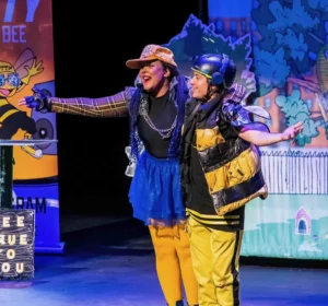 Two performers in bee-inspired costumes stand together on a colorful stage set with illustrated scenery and props during a children's theater production.