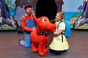 Two performers interact with Clifford the Big Red Dog on a colorful theater stage.