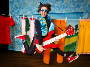 Performer wearing colorful stage attire and sunglasses leaps while holding an oversized red guitar in front of a bright storybook-style set with clothes hanging on a clothesline.