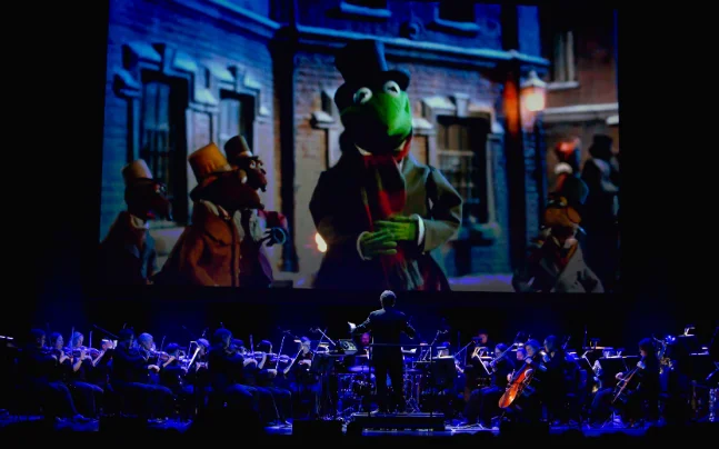 Disney’s The Muppet Christmas Carol Live in Concert featuring a live orchestra performing the film score beneath a large screen showing scenes from the holiday classic, including Kermit the Frog, Gonzo, and other Muppet characters.