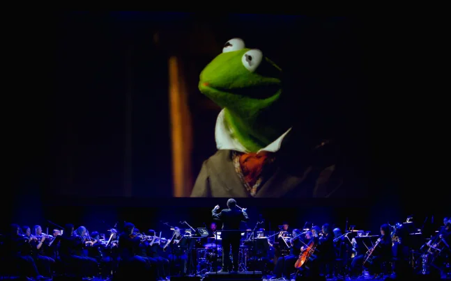 Disney’s The Muppet Christmas Carol Live in Concert featuring a live orchestra performing the film score beneath a large screen showing scenes from the holiday classic, including Kermit the Frog.