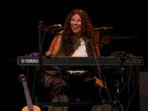 Performer at a Yamaha keyboard onstage with microphones, a tablet and a guitar nearby.
