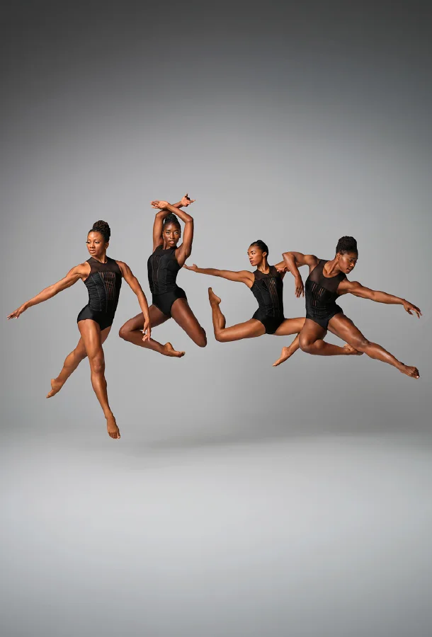 Four dancers in black performance attire performing dynamic jumps against a gray background.