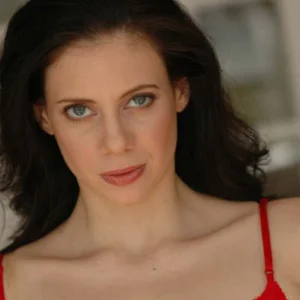 Professional headshot of Ashley Bachner wearing a red sleeveless top, looking toward the camera against a softly blurred indoor background.