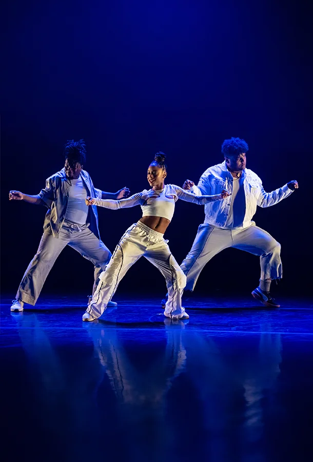 Three dancers in white costumes perform on a blue-lit stage in a Camille A. Brown & Dancers production.