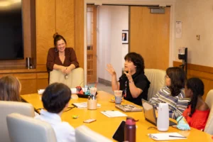 A small group of participants sit around a conference table during a discussion session. One participant gestures while speaking as others listen. A facilitator ands at the end of the table in a wood-paneled meeting room with open doors in the background.