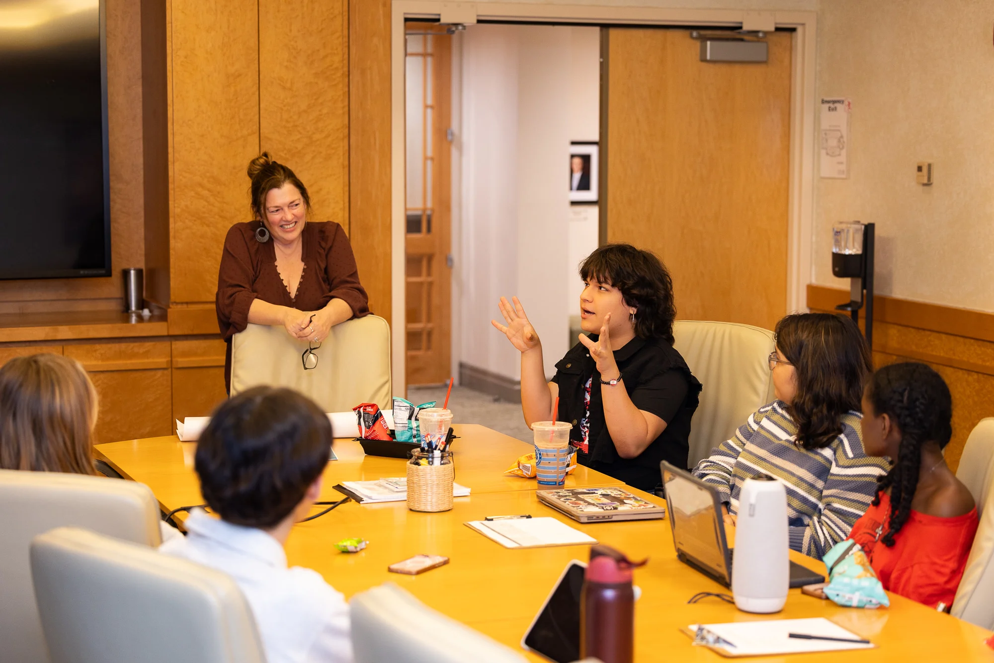 Changemakers Fall 2025 A small group of participants sit around a conference table during a discussion session. One participant gestures while speaking as others listen. A facilitator ands at the end of the table in a wood-paneled meeting room with open doors in the background.