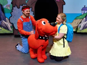 Two performers interact with Clifford the Big Red Dog on a colorful theater stage.