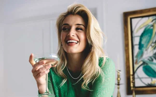 Daphne Oz seated indoors wearing a green dress and holding a glass.