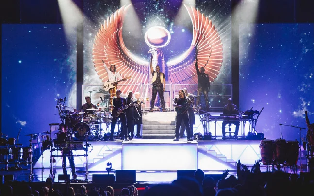 Earth, Wind & Fire live concert performance featuring the legendary band on a multi-level stage with dramatic lighting, a signature winged backdrop, and a full ensemble of musicians. Dynamic concert photography showcasing the group's iconic stage production at the Kravis Center.