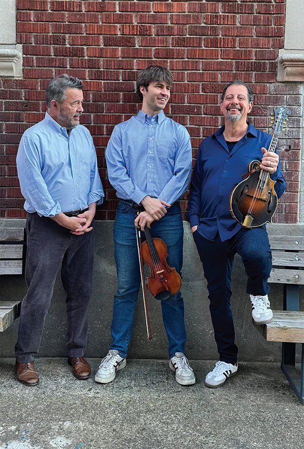 Members of Edgar, George & Mike pose outdoors with a violin and mandolin in a promotional portrait.