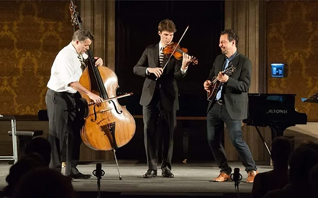 Live performance by the Edgar, George & Mike acoustic ensemble featuring three musicians on stage playing double bass, violin, and mandolin. The trio performs in an elegant concert hall setting, showcasing virtuosic string musicianship rooted in folk, Celtic, bluegrass, and American roots music traditions.