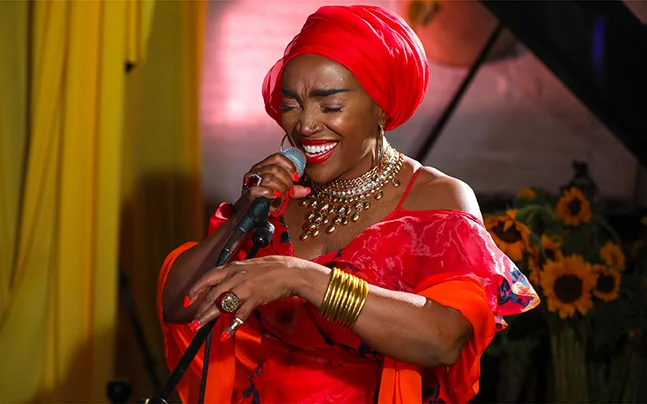 Performer singing into a microphone during a live concert while wearing a vibrant red outfit and headwrap.