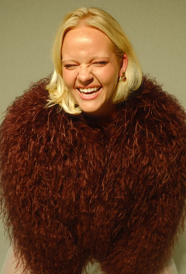 Portrait of vocalist Emma Nissen laughing and wearing a brown textured coat against a neutral background.