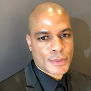 Headshot of Herman Payne wearing a black suit and black shirt, photographed against a neutral gray background.