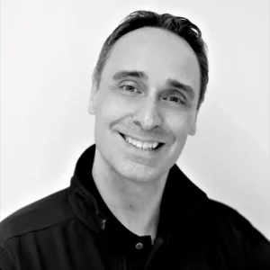 Black-and-white portrait of Jean-Hugues Feray wearing a dark jacket, smiling toward the camera against a light background.