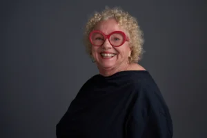 Studio portrait of a person with short curly hair wearing large round red eyeglasses and a dark top. The person is posed against a dark gray background.