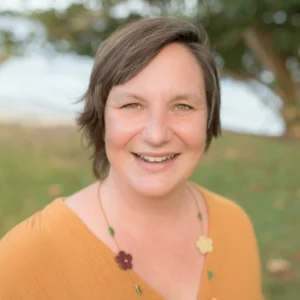 Outdoor portrait of Kori Epps wearing a mustard-colored top and floral necklace, standing in a grassy area with trees in the background.