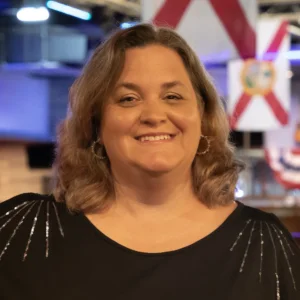 Portrait of Kristi Moynihan wearing a black top, photographed indoors with decorative Florida flags and soft lighting in the background.