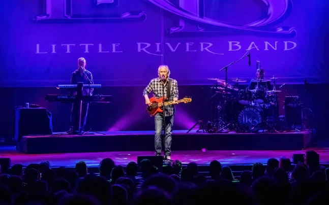 Little River Band performing live on stage with guitar, keyboards, and drums under purple concert lighting.