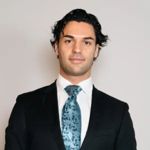 Professional headshot of Manuel Favilla wearing a dark suit, white dress shirt and patterned blue tie against a light gray background.