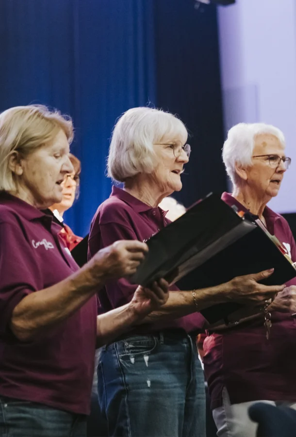 Community choir members singing on stage while holding music folders during a live performance.