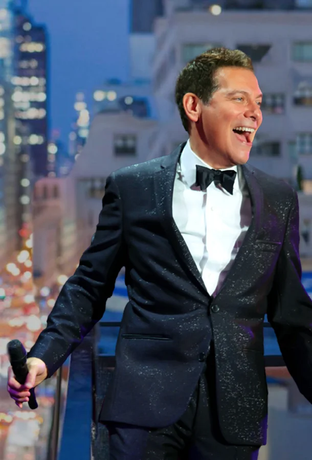 Performer in a black tuxedo holding a microphone with a city skyline in the background.