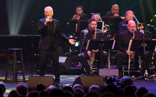 Live concert image featuring a vocalist performing on stage with a big band orchestra. The singer stands at the front of the stage holding a microphone, while seated musicians perform with saxophones, brass instruments, and music stands behind. Colorful stage lighting illuminates the performers and audience.