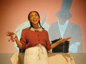 A performer in period-style clothing stands beside a table onstage, with projected imagery and a figure in a top hat visible in the background as part of a theatrical production.