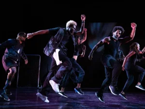 A group of dancers dressed in black leap across a stage in a synchronized performance under dramatic theatrical lighting.